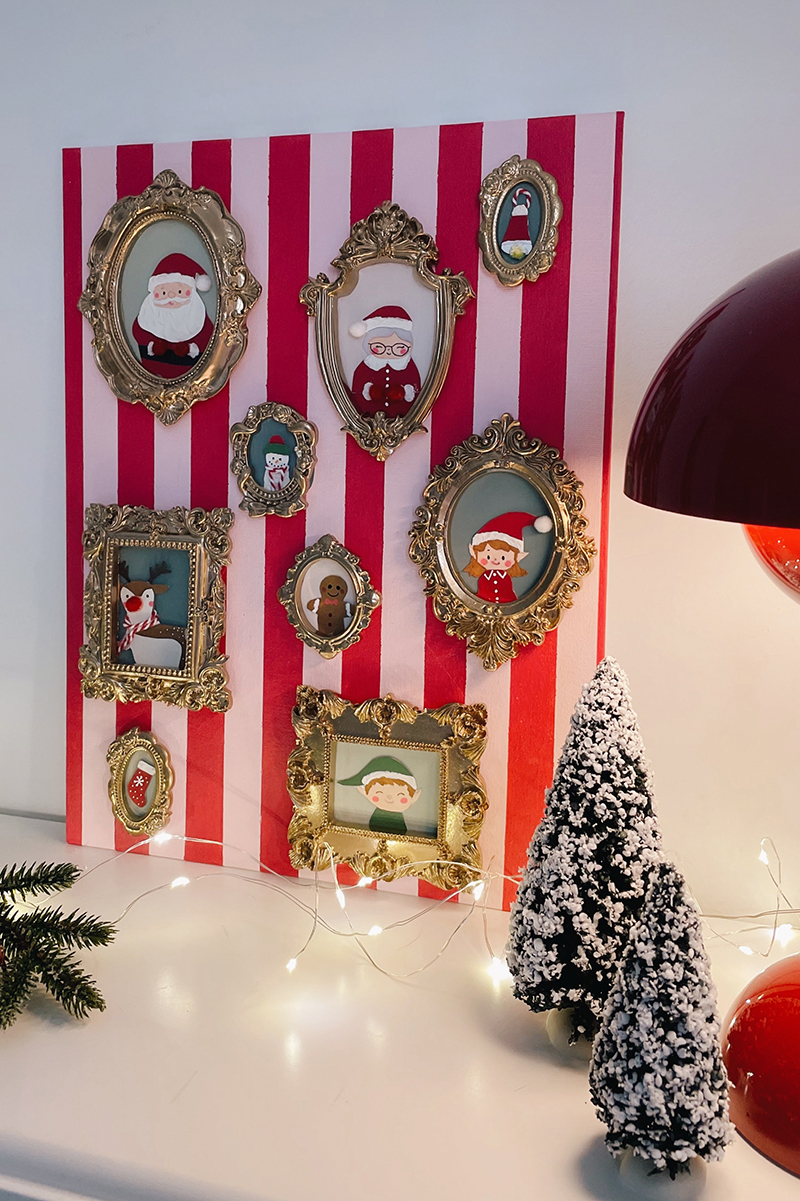 Mur décoratif de Noël avec mini-cadres dorés contenant des personnages illustrés sur un fond rayé rose et rouge, entouré de sapins enneigés et de guirlandes lumineuses.