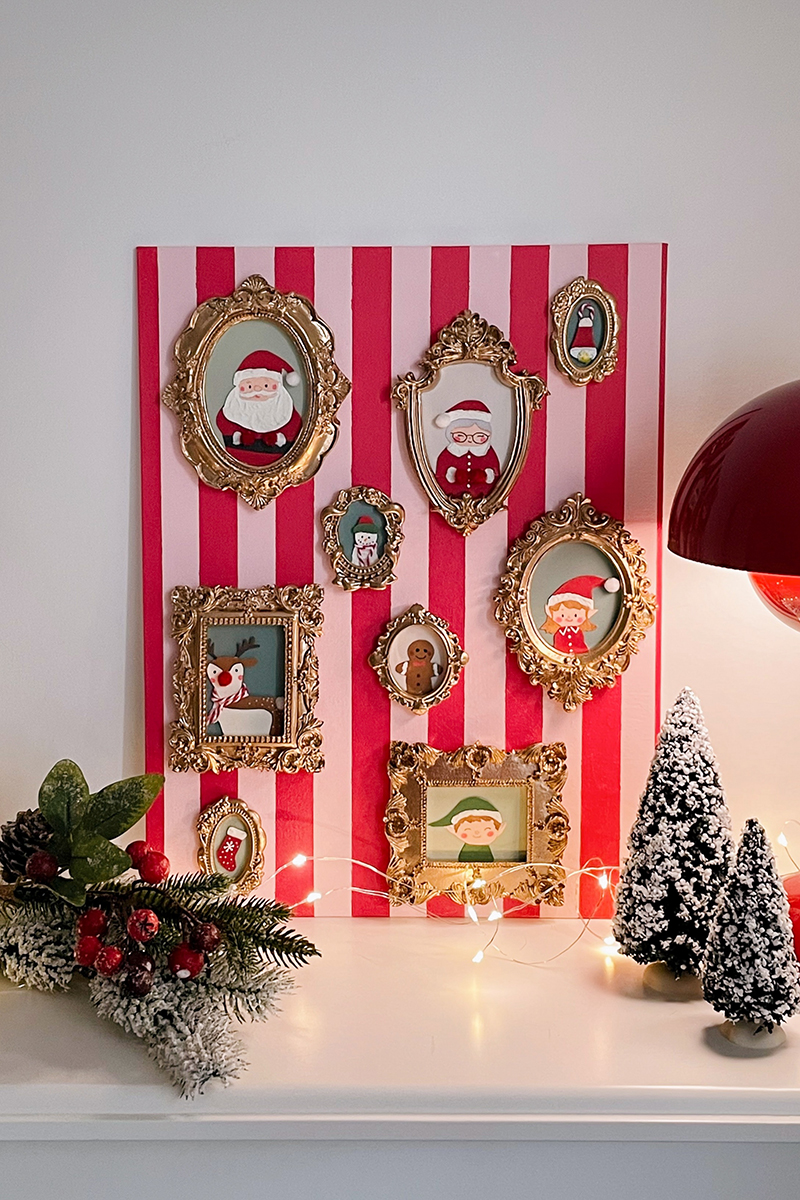 Composition murale de Noël avec cadres dorés et portraits illustrés sur fond rayé rouge et rose, décorée de sapins enneigés et de guirlandes lumineuses.