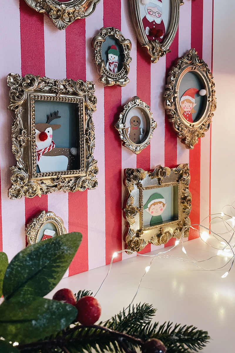 Vue latérale d’un mur de cadres dorés affichant des portraits de Noël illustrés, avec un renne, un biscuit et un lutin, sur fond rayé rose et rouge.